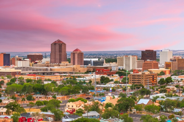 Cityscape of Albuquerque at sunset, showcasing buildings against a colorful sky with greenery in the foreground.