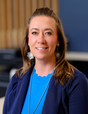 A woman with long brown hair, wearing a blue top and navy cardigan, smiles in an office setting.