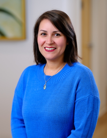 A woman with shoulder-length dark hair smiles while wearing a blue sweater in an indoor setting.