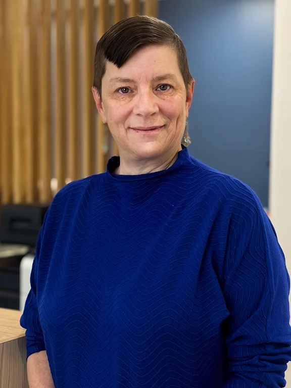 A person with short hair wearing a blue sweater stands in an office setting, smiling at the camera.