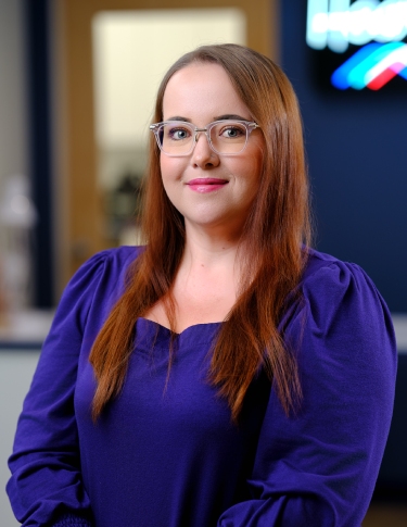 A woman with long brown hair in a purple blouse smiles at the camera, posing in an office setting.