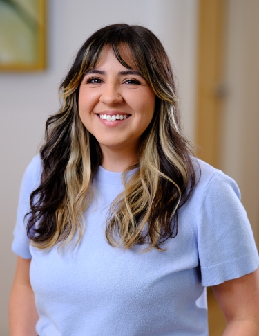 A woman with long, wavy hair featuring highlights, smiling while wearing a light blue shirt in a well-lit indoor setting.