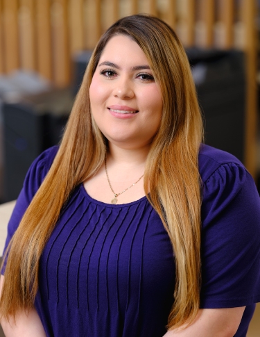 A woman with long, straight hair smiles while wearing a purple top, posing for a portrait in a well-lit setting.