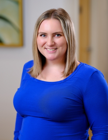 A woman with shoulder-length blonde hair smiles while wearing a blue long-sleeve shirt. A softly lit background is blurred.