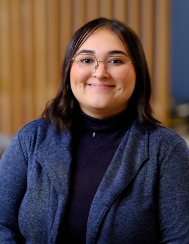 A young person with glasses and dark hair smiles, wearing a navy cardigan over a black turtleneck, against a blurred background.