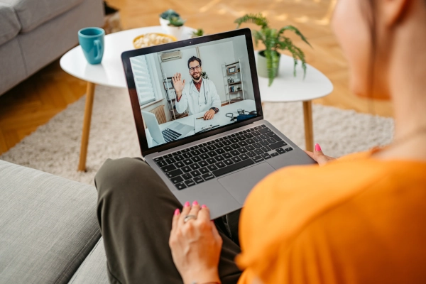 A person sitting on a couch is having a telehealth consultation on a laptop with a doctor visible on the screen.