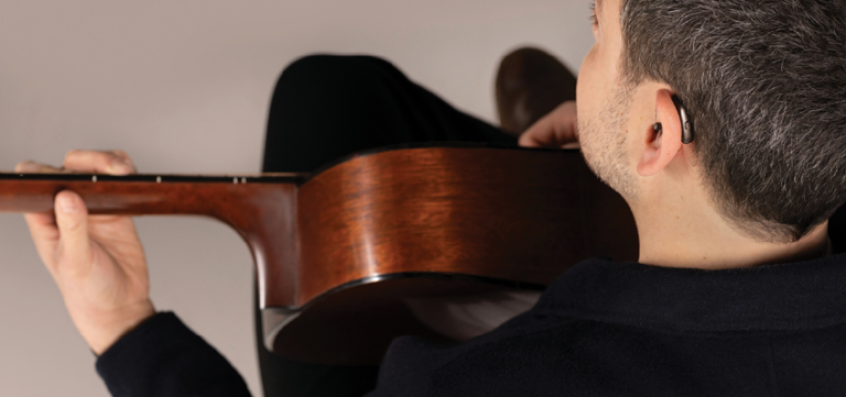 A man wearing a hearing aid plays an acoustic guitar