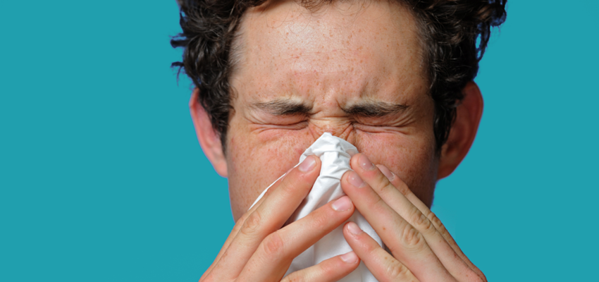 A close-up of a person with curly hair sneezing into a white tissue against a bright teal background