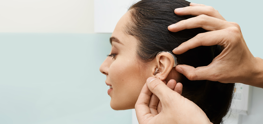 A professional's hands carefully adjusting a behind-the-ear hearing aid on a woman's ear during a clinical fitting