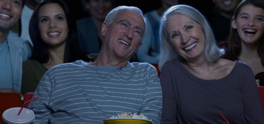 An older couple smiling and laughing in a movie theater with popcorn and a drink, surrounded by a cheerful audience