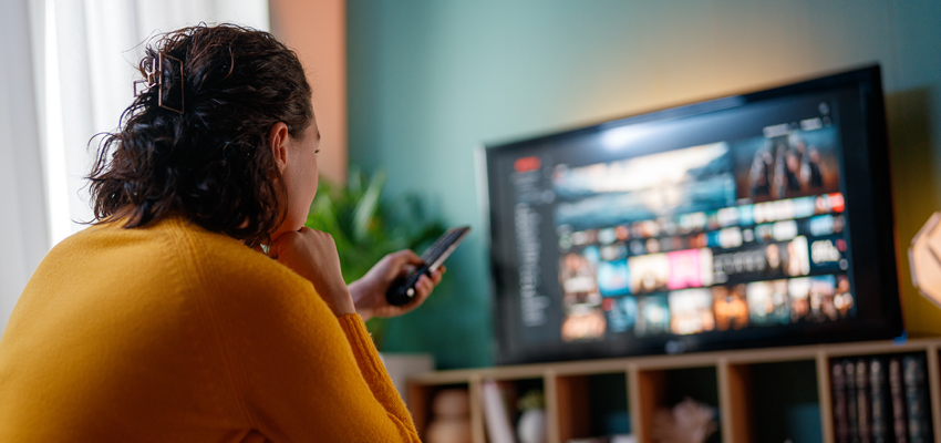 A person in a yellow sweater sits on a couch, holding a remote control and browsing a streaming menu on a television screen