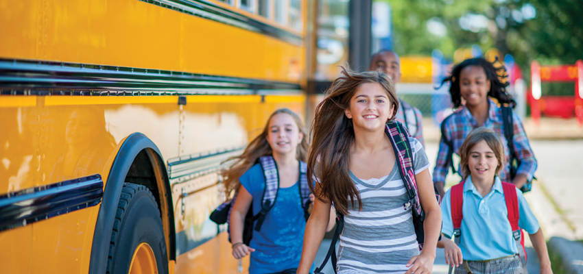 Smiling students wearing backpacks run excitedly away from a yellow school bus