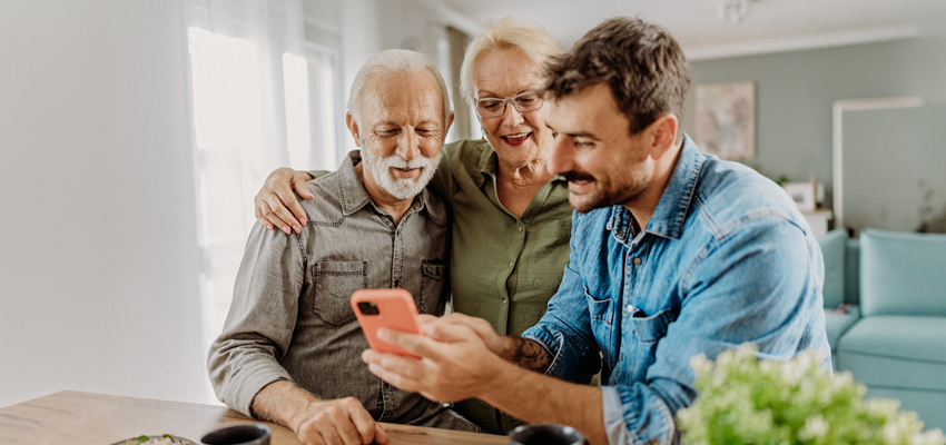 A young man shows a smartphone screen to a smiling older couple leaning in closely together